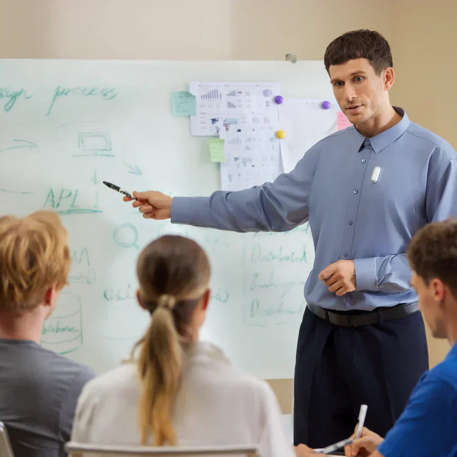 A man in a blue shirt points at a whiteboard while wearing a silver Plaud wearable AI recorder to capture the meeting.