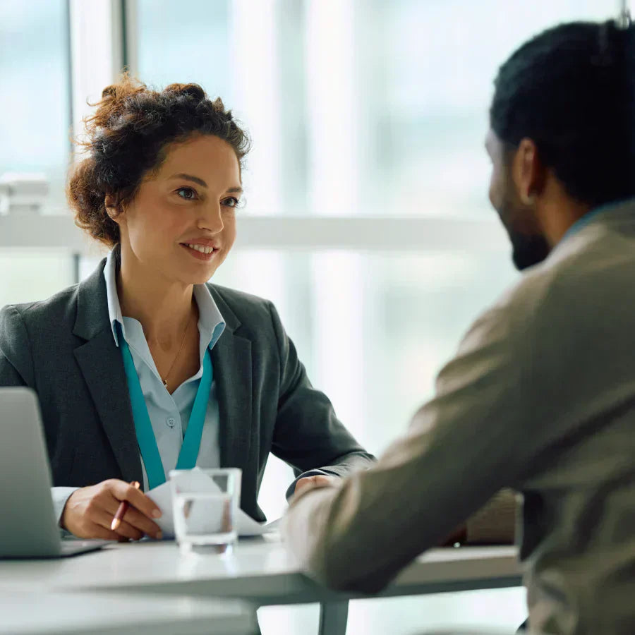 A professional woman wearing a Plaud wearable AI recorder on a teal lanyard speaks with a colleague in a bright office.