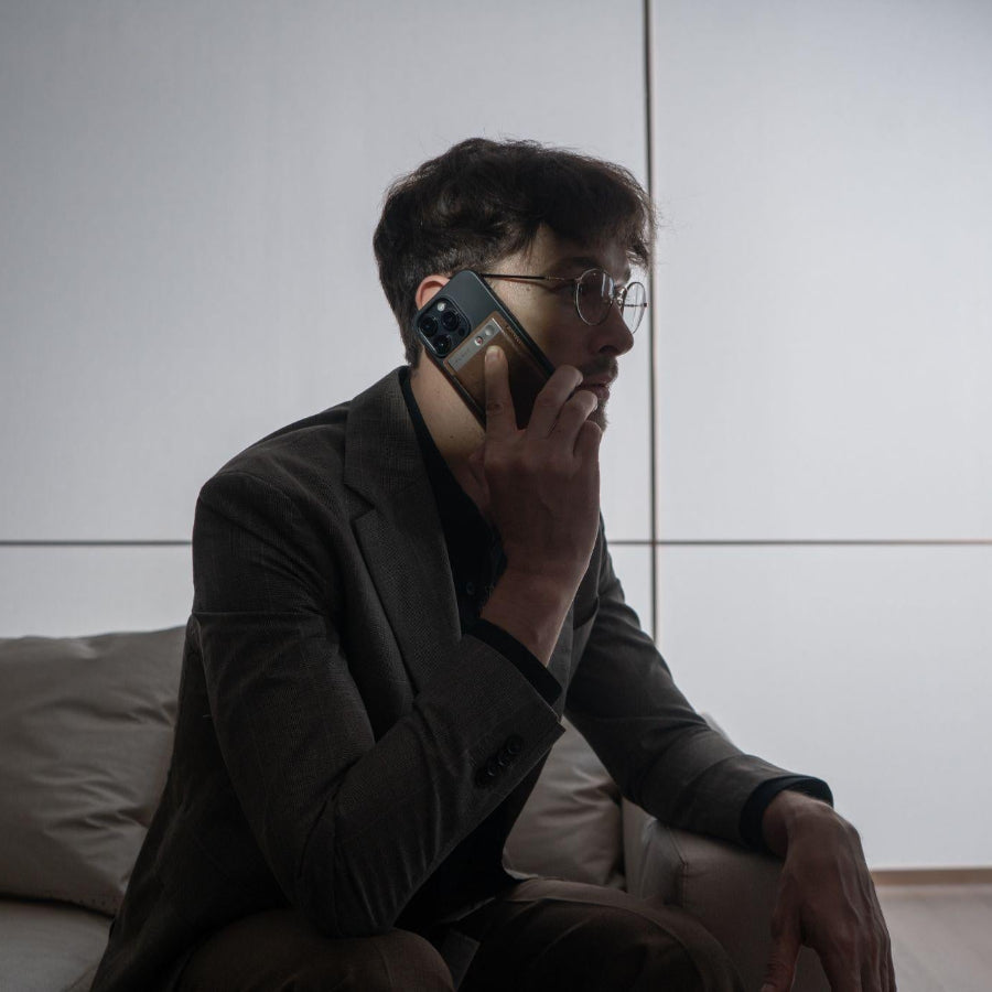 A man in a suit jacket holds a smartphone to his ear with a starlight Plaud Note AI voice recorder attached to the back.