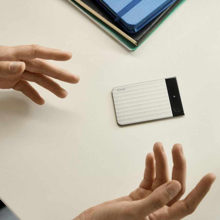 A silver Plaud Note Pro lies on a white desk between a person's gesturing hands and a stack of colorful notebooks.