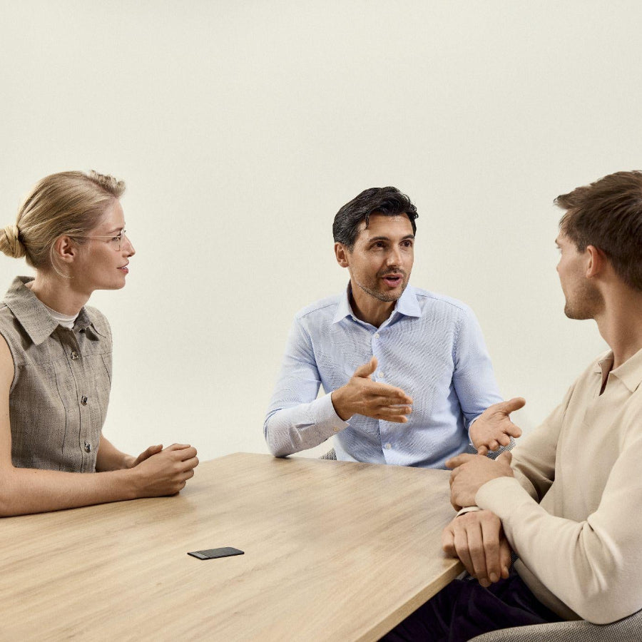 Three professionals sit at a wooden table having a conversation while a black Plaud AI voice recorder captures their meeting.