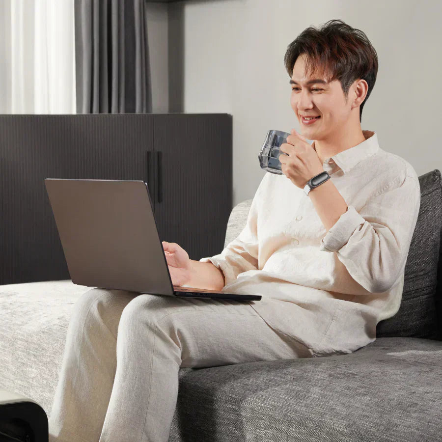 A man in a beige linen shirt smiles while using a laptop and drinking from a glass, wearing a Plaud wearable AI recorder on his wrist.
