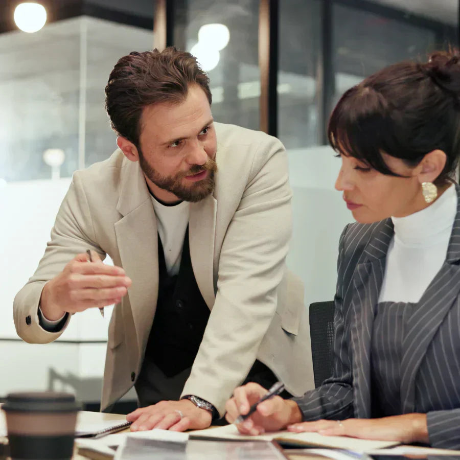 A man in a light blazer leans over a desk to discuss notes with a woman in a pinstripe suit during a meeting.
