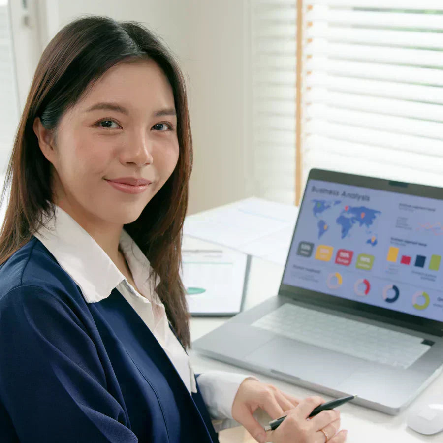 A woman in a navy blazer smiles while sitting at a desk with a laptop displaying a business analysis dashboard.