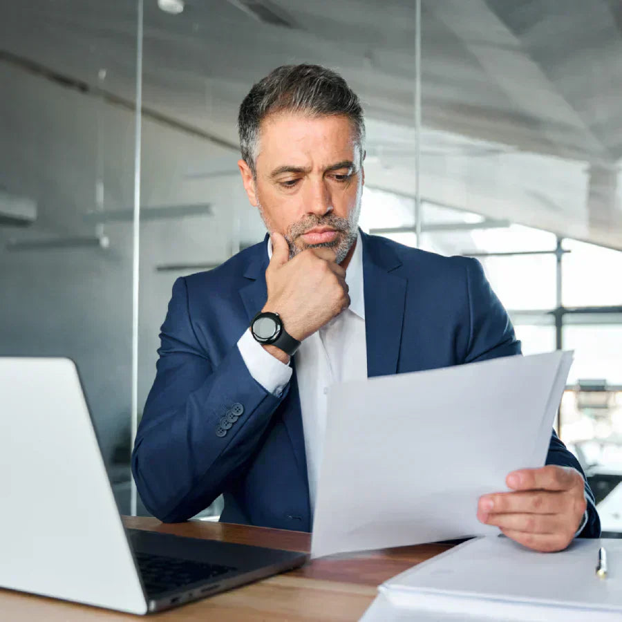 A professional man in a blue suit sits at a desk, thoughtfully reviewing a document while resting his chin on his hand.