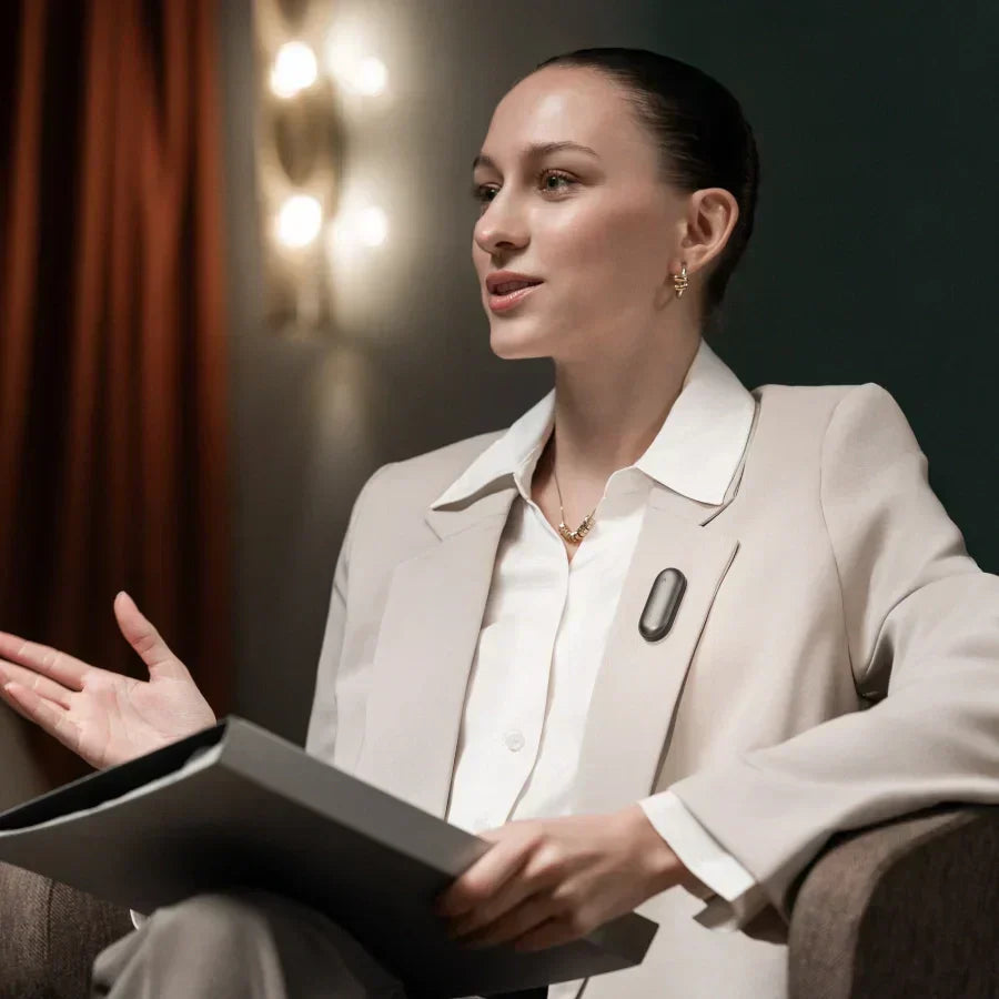 A professional woman in a beige blazer speaks while wearing a dark Plaud wearable AI recorder on her lapel.