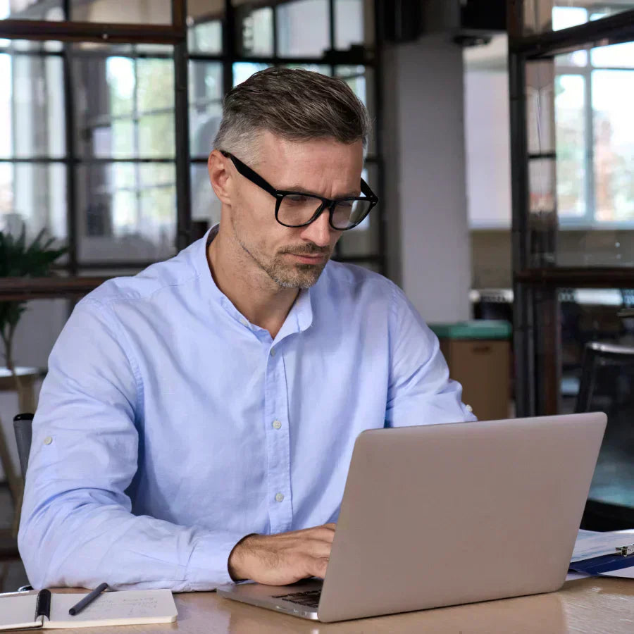 A professional man in a light blue shirt and glasses focuses on his laptop while working at a desk in an office.