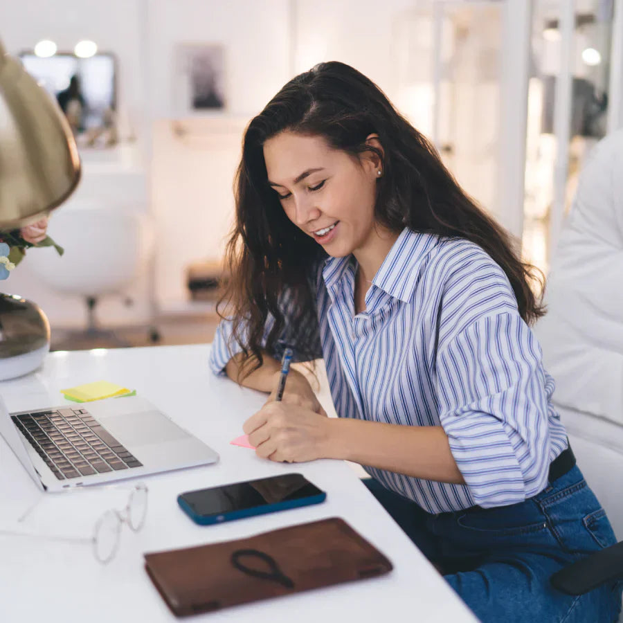 A woman in a striped shirt sits at a white desk, smiling as she writes on a pink sticky note next to her laptop.