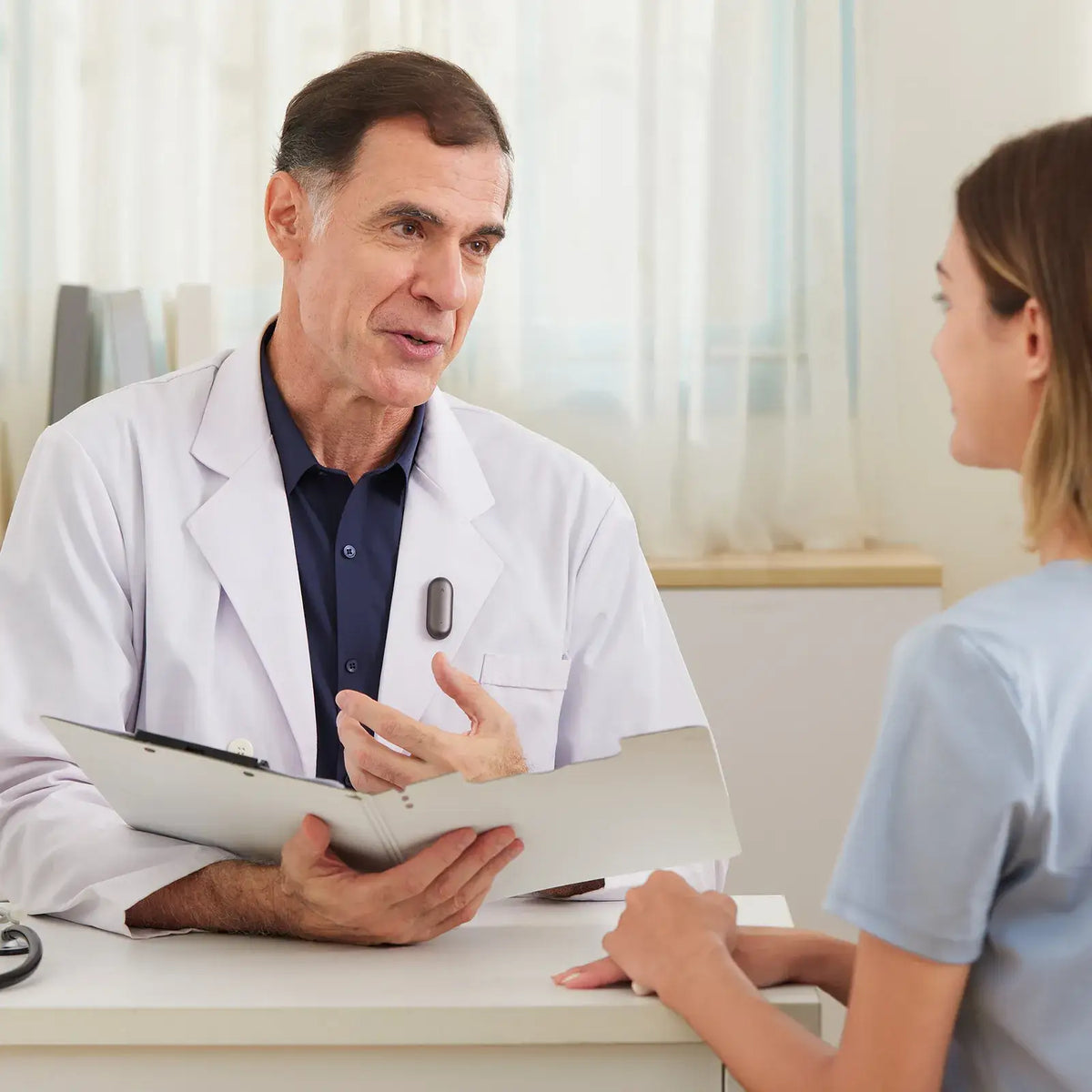 A doctor in a white coat speaks with a patient while wearing a Plaud wearable AI recorder on his lapel.