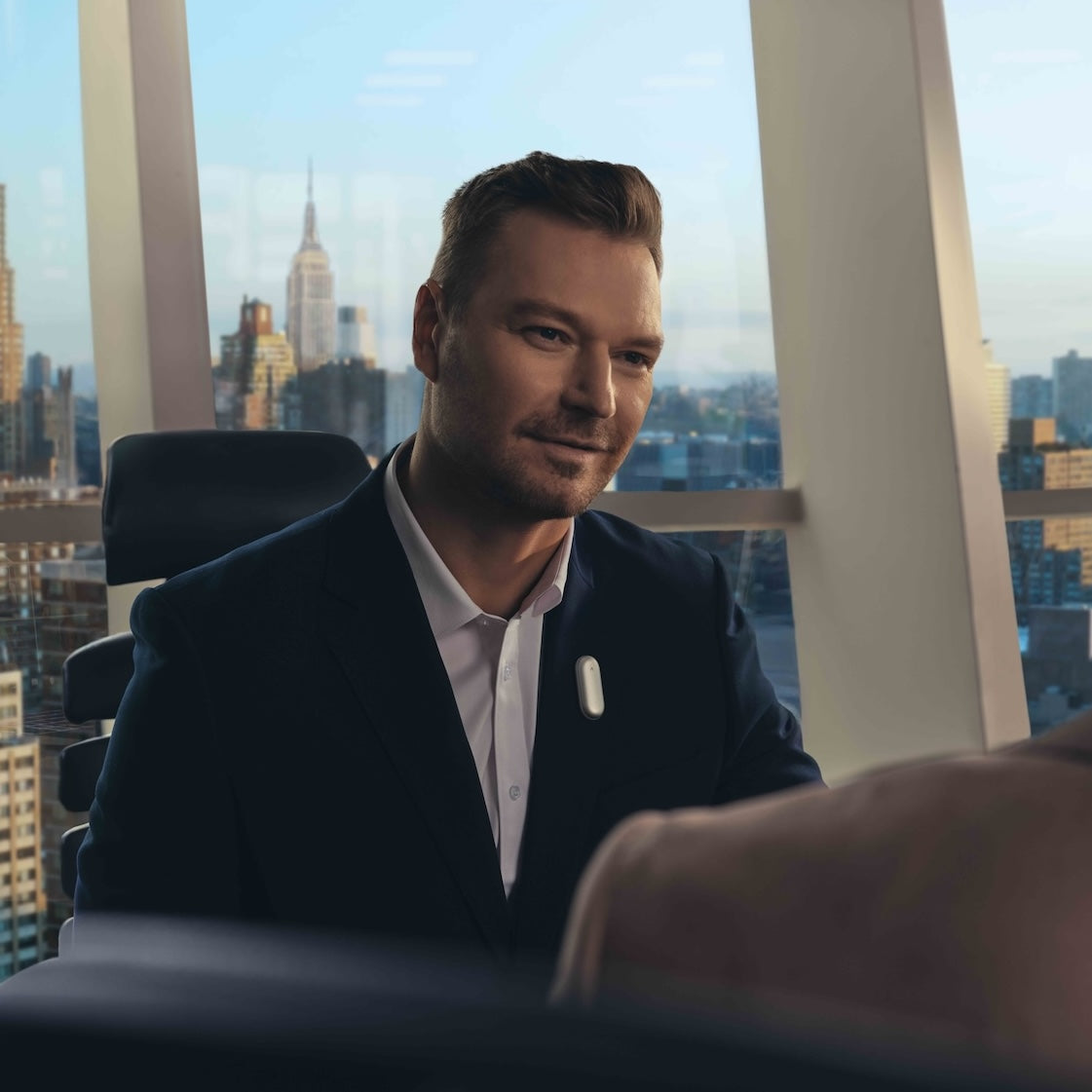 A man in a navy suit sits in a high-rise office with a city view, wearing a white Plaud wearable AI recorder on his lapel.