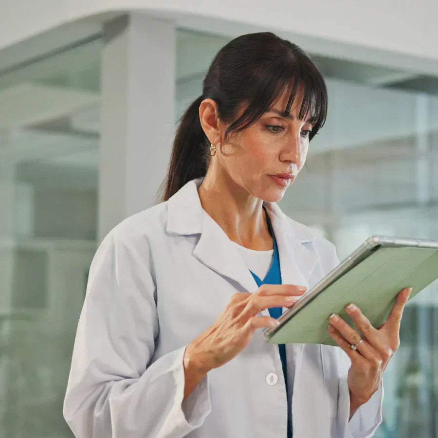 A healthcare professional wearing a white lab coat and blue scrubs uses a tablet in a light green case within a bright clinic.