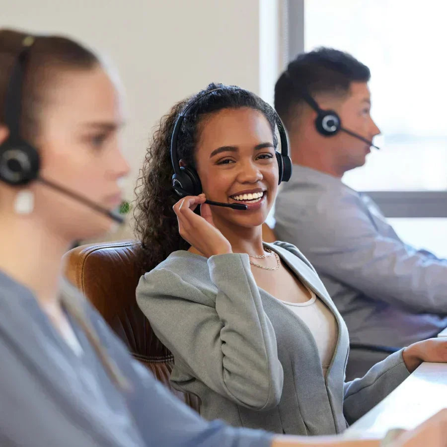 A smiling professional woman wearing a black headset works in a bright office alongside two colleagues.